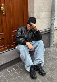 Personne portant un bonnet noir, des lunettes, une veste en cuir noire, un jean bleu clair et des baskets noires assise sur une marche en béton près d'une porte en bois.