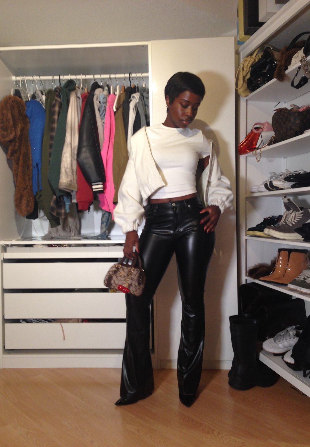 Vrouw in een witte cropped top en zwarte leren broek staat in een kast, terwijl ze een kleine gepatternde handtas vasthoudt, omringd door hangende kleding en schoenen.