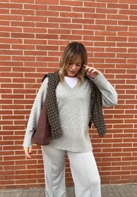 Kvinna som står mot en röd tegelvägg iförd en grå tröja, vita byxor, en rutigt jacka över axlarna och håller en brun väska.