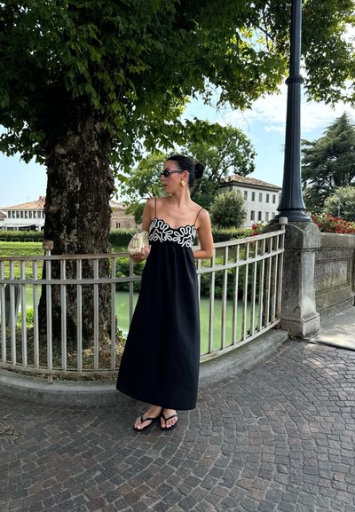 Vestido maxi negro con un patrón abstracto blanco en el torso. La modelo sostiene un bolso pequeño y lleva chanclas negras y gafas de sol.