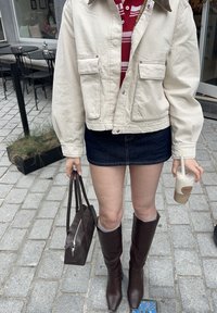 Kremfarget canvasjakke med to lommer, rød og hvit stripete skjorte under, mørk denim miniskjørt, brune høye støvler, holder en drink.