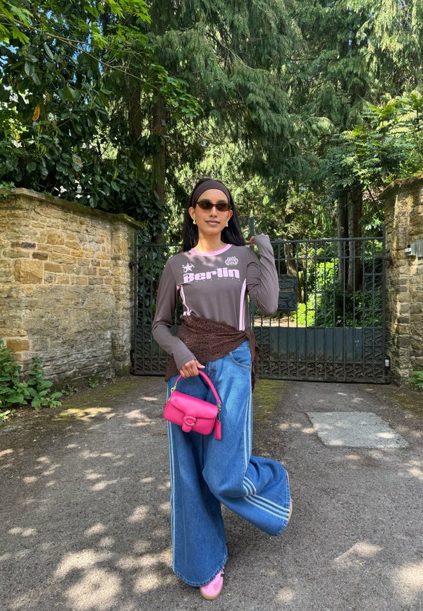 Femme portant des lunettes de soleil, un chemisier marron à manches longues avec l'inscription « Berlin », un jean large bleu, des chaussures roses, et tenant un petit sac à main rose vif à l'extérieur.