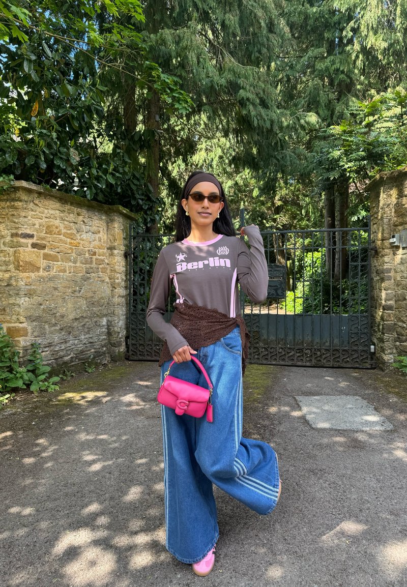 Femme portant des lunettes de soleil, une chemise marron à manches longues avec le mot "Berlin", un jean large bleu, des chaussures roses, et tenant un petit sac à main rose vif en extérieur.