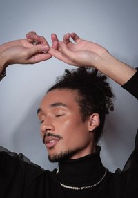 Personne portant un pull noir à col roulé avec des manches transparentes, orné d'une chaîne en argent et d'un collier de perles, les mains levées dans une pose gracieuse.