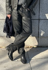 Fekete bőrdzseki és nadrág, alatta szürke pulóver, sötét sállal a kézben. Négyzet alakú orrú fekete csizmák, egy texturált kőfalnak támaszkodva.