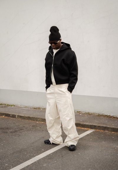 Sudadera negra con cremallera y corte oversized, suéter crema debajo, pantalones anchos blancos y zapatos negros. De pie contra una pared lisa.