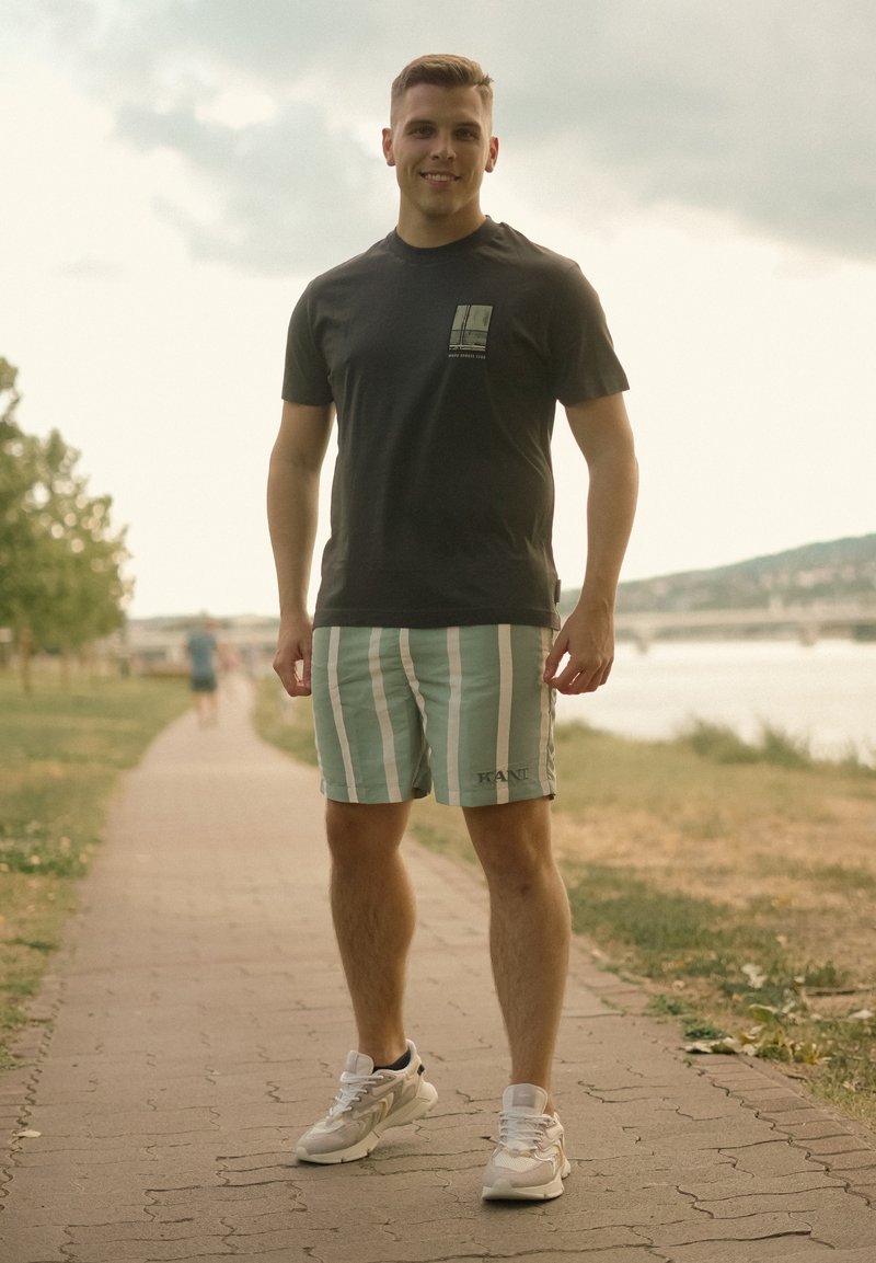 Jeune homme souriant, debout sur un sentier près d'une rivière, portant un t-shirt noir, un short à rayures et des baskets blanches.