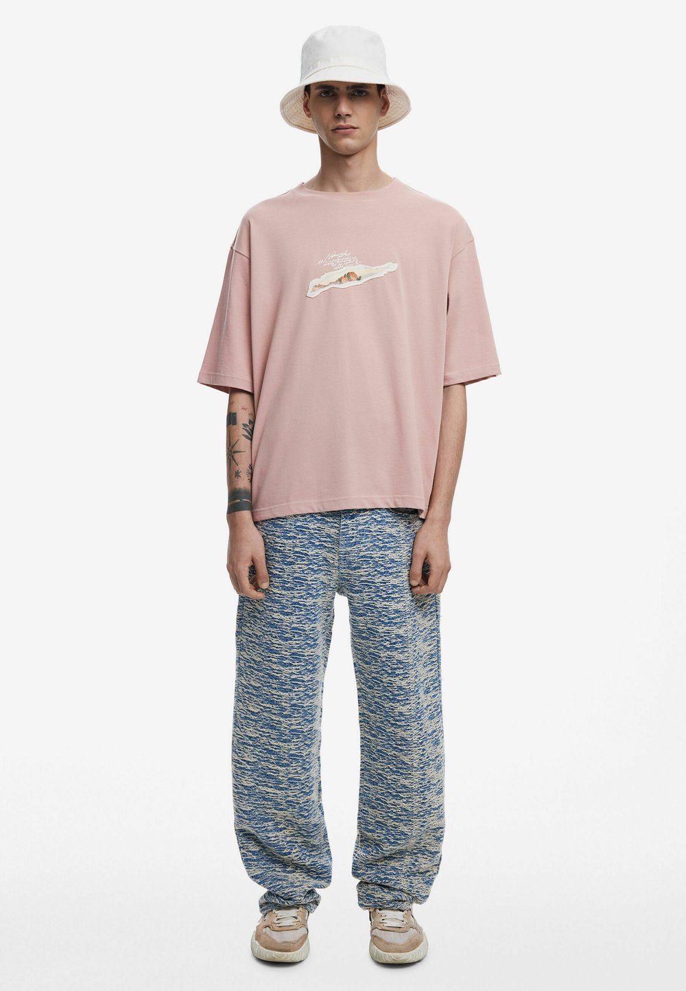 Tricou roz cu mânecă scurtă și detaliu grafic, asortat cu pantaloni texturați albaștri și adidași bej. O pălărie albă tip găleată completează look-ul.