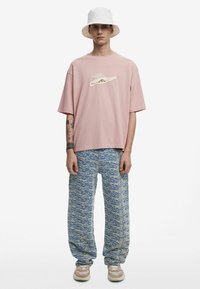 Tricou roz cu mânecă scurtă și detaliu grafic, asortat cu pantaloni texturați albaștri și adidași bej. O pălărie albă tip găleată completează look-ul.