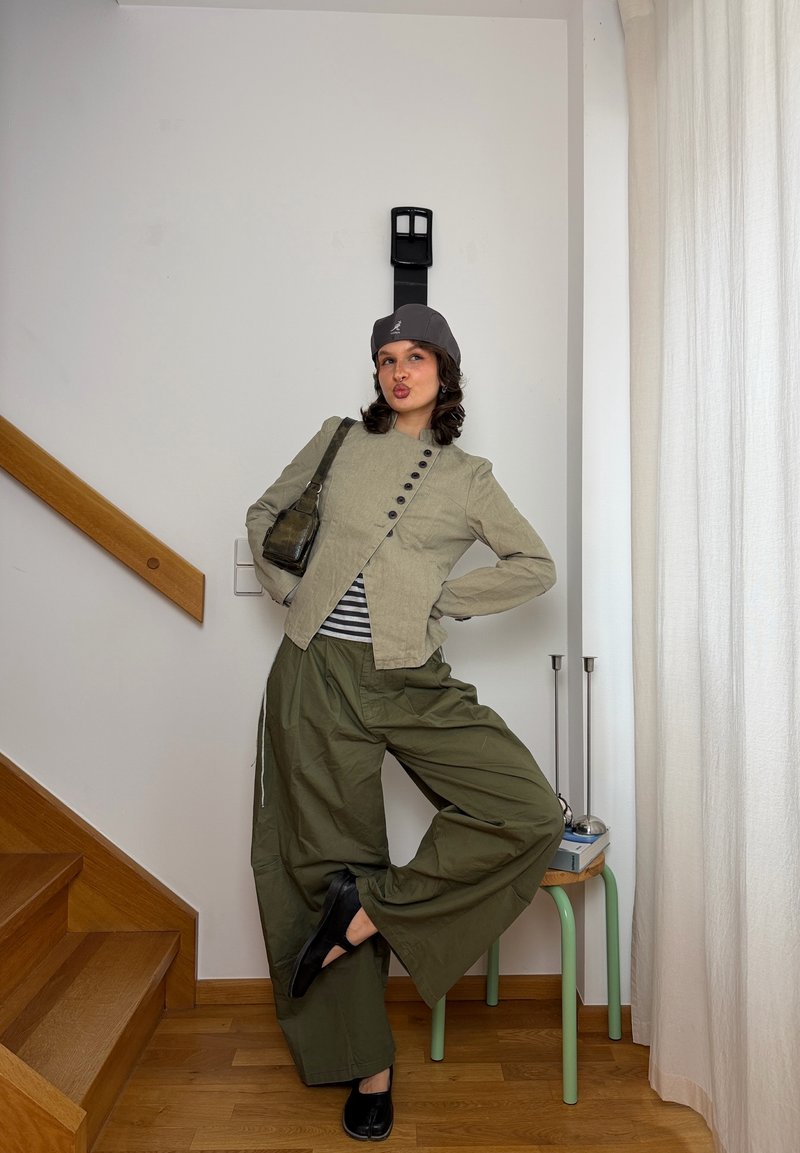 Joven mujer con pantalones anchos verdes, chaqueta beige, gorra gris y zapatos negros posa con una pierna levantada en una habitación con escaleras de madera y cortinas blancas.