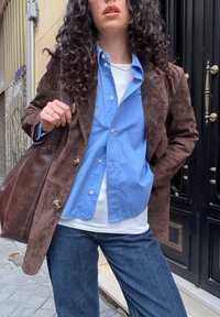 Personne aux cheveux bouclés portant une veste en daim marron, une chemise bleue boutonnée sur un haut blanc, un jean bleu, et portant un grand sac en cuir marron.