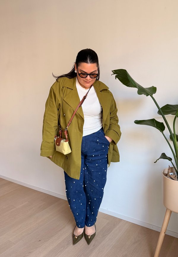 Žena u maslinasto zelenoj jakni, bijelom topu, plavim hlačama s bijelim točkicama, nosi malu bež torbu preko ramena, stoji pored biljke u tegli.