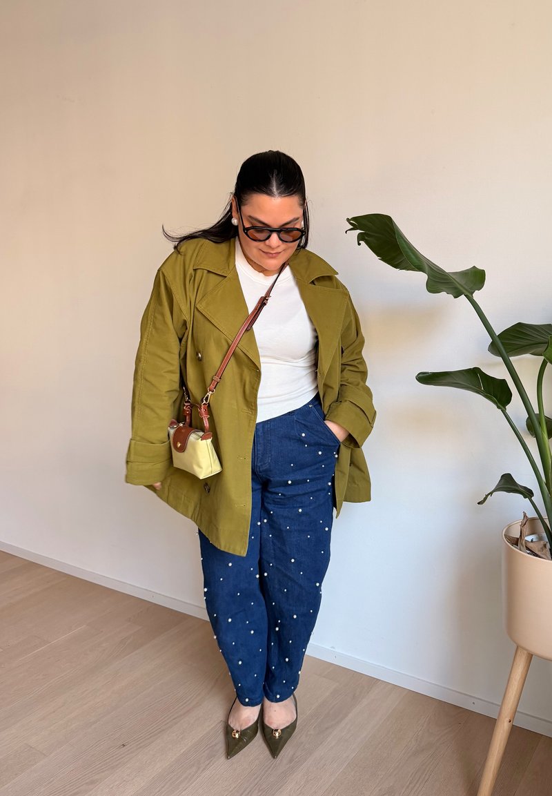 Moteris, dėvinti alyvuogių žalią striukę, baltą palaidinę, mėlynas kelnes su baltais taškeliais, nešioja mažą smėlio spalvos rankinę per petį, stovi šalia vazoninės gėlės.