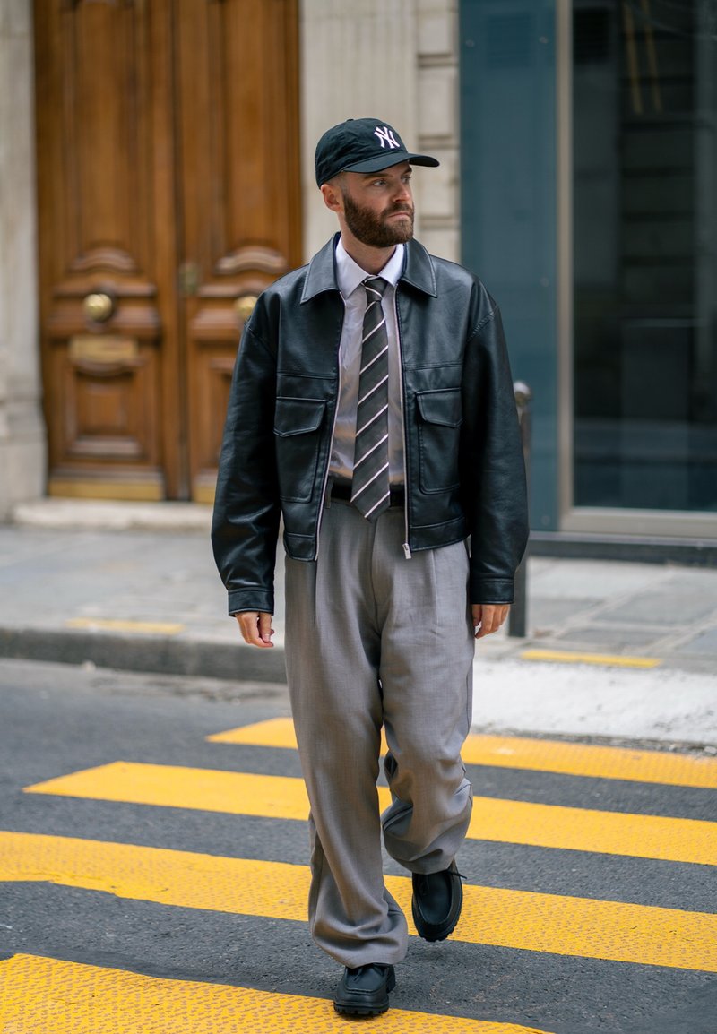 Jaqueta de couro preta, camisa branca e gravata listrada, calças cinzas, boné preto com logotipo e sapatos pretos, caminhando numa rua com faixas amarelas.
