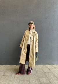 Beige blank trenchcoat, krem genser, brune joggebukser med hvite striper, burgunder sneakers, grønn caps og liten grønn håndveske.