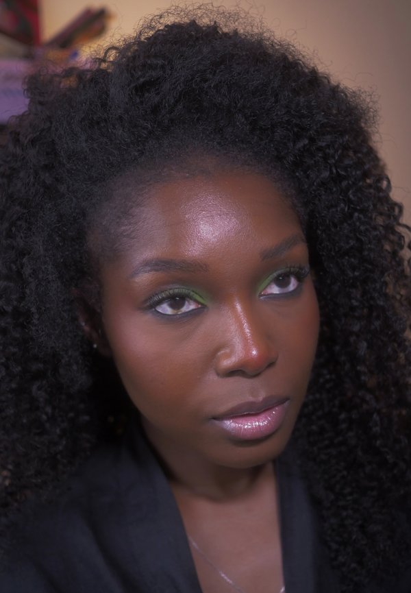 Femme aux cheveux foncés bouclés, fard à paupières vert et lèvres brillantes regardant légèrement vers le haut et sur le côté sur un fond flou.