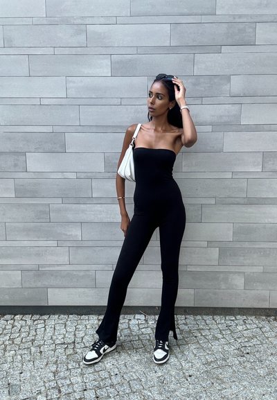 Mono negro sin tirantes con un diseño ajustado, combinado con un bolso blanco y zapatillas en blanco y negro. Fondo de pared gris texturizado.