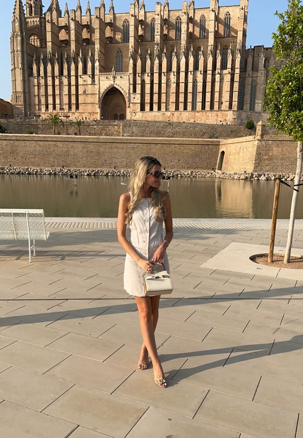 Světle béžové šaty bez rukávů s detaily knoflíků, v kombinaci s malou bílou kabelkou a sandály na podpatku. Venku, blízko odrazového vodního tělesa.