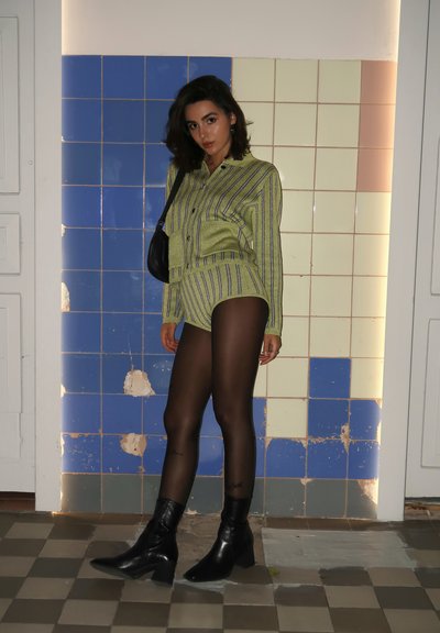 Conjunto de camisa y pantalones cortos a rayas verdes y azules, combinado con medias negras transparentes y botas negras hasta el tobillo. El fondo presenta paredes de azulejos en colores suaves.