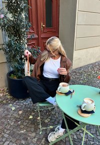Bruine leren jas over een witte shirt, gestreepte broek, zittend aan een groene tafel met twee mokken op houten onderzetters. Klinkers als achtergrond.