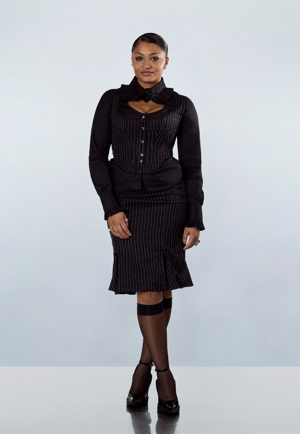 Γυναίκα όρθια, με κορσέ μαύρο με λεπτές ρίγες και φούστα, διάφανα μαύρα καλσόν μέχρι το γόνατο, μαύρα τακούνια, με τα χέρια χαλαρά στο πλάι.