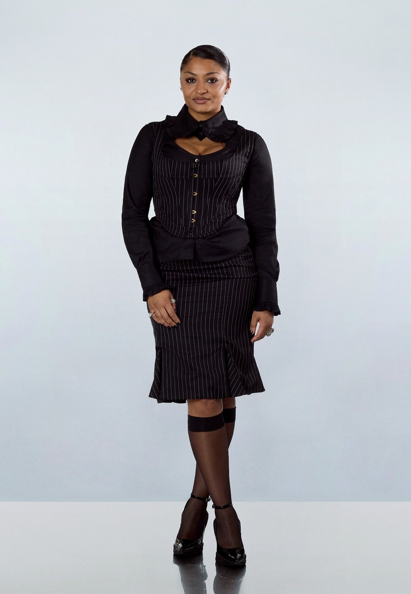 Vrouw staand, gekleed in een zwart korsetachtige top met krijtstreep en rok, doorschijnende zwarte kniehoge kousen, zwarte hakken, met handen ontspannen langs haar zij.