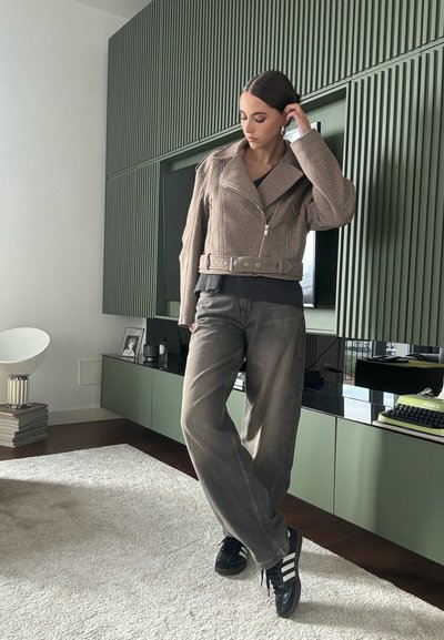 Chaqueta de moto marrón cortada con una cintura con cinturón, combinada con pantalones grises holgados y zapatillas negras, todo frente a un interior moderno.