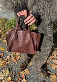 Geantă maro din piele cu două mânere scurte, textură netedă și formă structurată, ținută în mâini deasupra frunzelor căzute și a trotuarului.