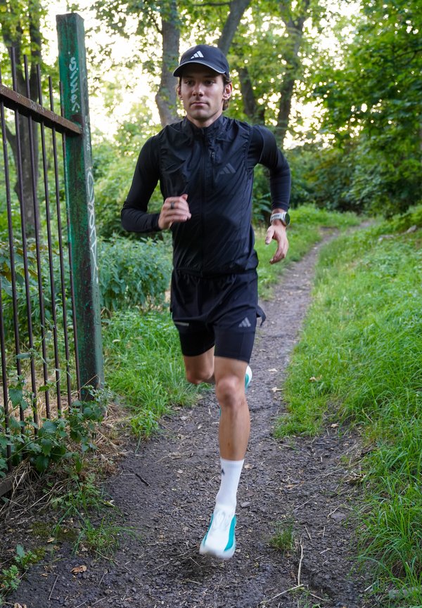Muškarac obučen u crnu sportsku odjeću i kapu trči zemljanim putem kroz zeleno šumsko područje blizu metalne ograde.