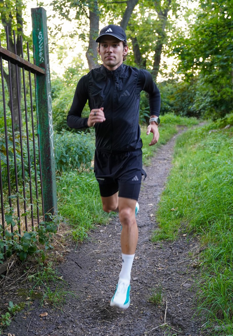 Homme portant des vêtements de sport noirs et une casquette, courant sur un sentier en terre à travers une zone forestière verte près d'une clôture métallique.