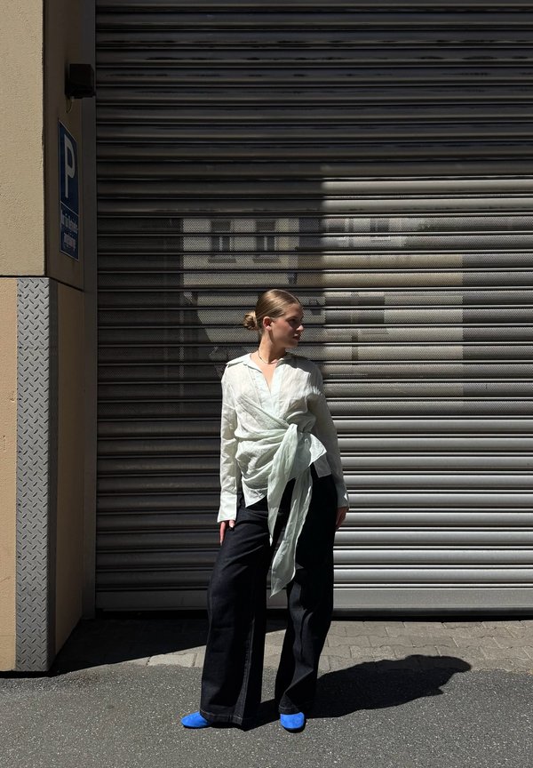 Femme en blouse blanche transparente avec un lien à la taille et un pantalon noir large, debout sur le trottoir devant une porte métallique à volet roulant, projetant une ombre.