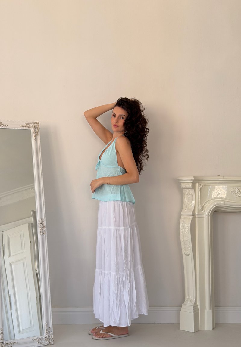 Donna con lunghi capelli ricci che indossa un top azzurro senza maniche e una gonna bianca, posa accanto a un grande specchio ornato e un camino decorativo bianco.