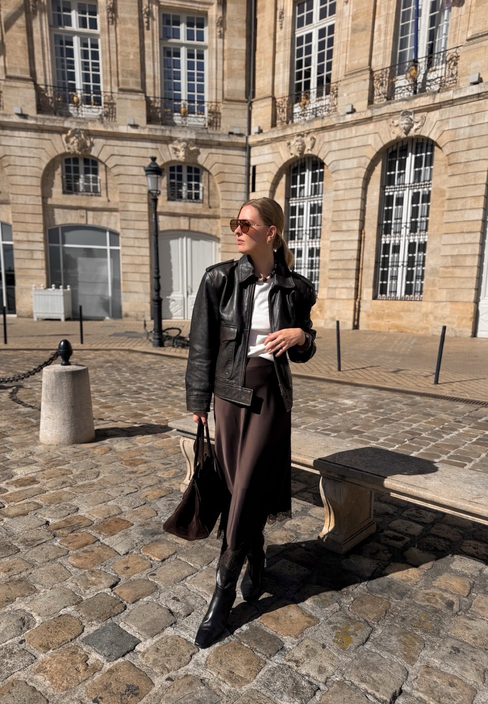 Black leather jacket over a white shirt, brown skirt with lace hem, and black heeled boots. Holding a brown bag, standing on cobblestone street.