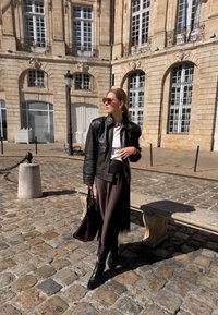 Black leather jacket over a white shirt, brown skirt with lace hem, and black heeled boots. Holding a brown bag, standing on cobblestone street.
