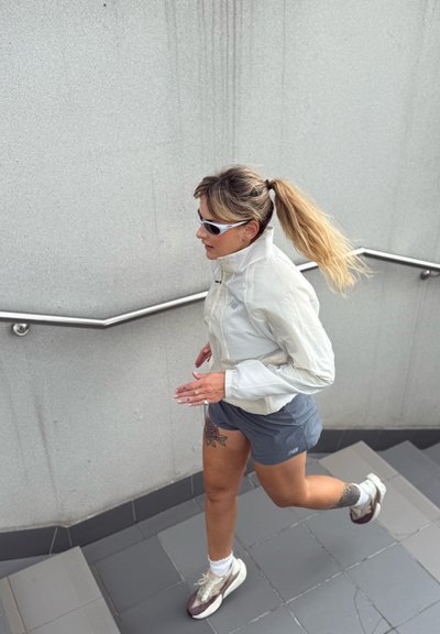 Mujer corriendo por unas escaleras interiores, vestida con una chaqueta blanca, pantalones cortos grises, gafas de sol y zapatillas, con el cabello recogido en una coleta y tatuajes visibles.