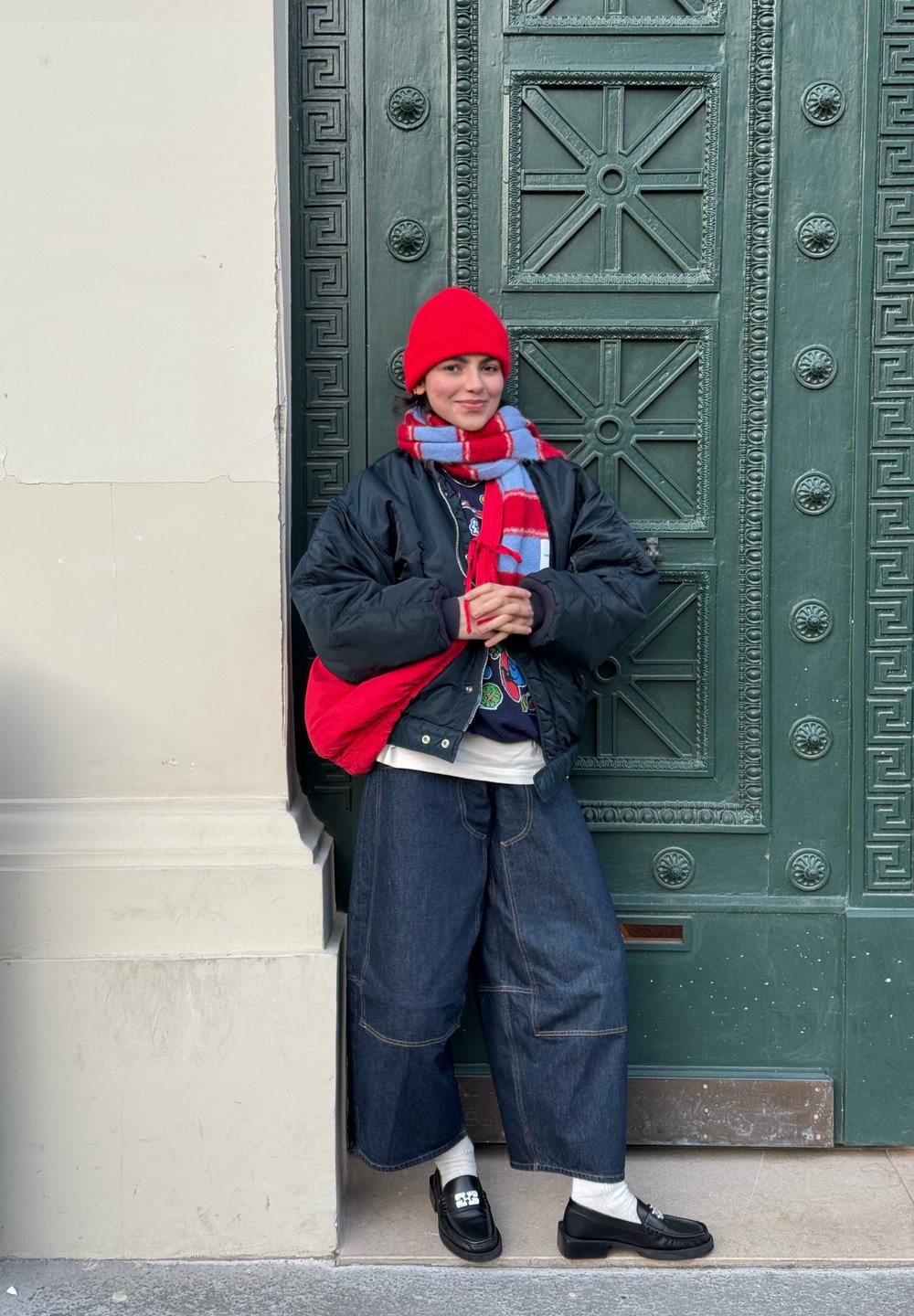 Personne portant un bonnet rouge en tricot et une écharpe assortie, appuyée contre une porte verte ornée, vêtue d'une veste noire, d'un pantalon large en jean, de chaussures noires, et tenant un sac rouge.