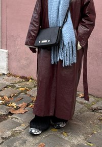 Personne portant un long manteau en cuir bordeaux, une grosse écharpe bleu pâle, un sac noir porté en bandoulière et des baskets argentées sur une rue pavée jonchée de feuilles mortes.