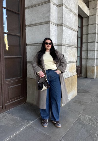 Abrigo largo y sobredimensionado de color marrón sobre un suéter claro, con vaqueros anchos de color azul y zapatillas grises. Bolso en blanco y negro con banda de hombro.