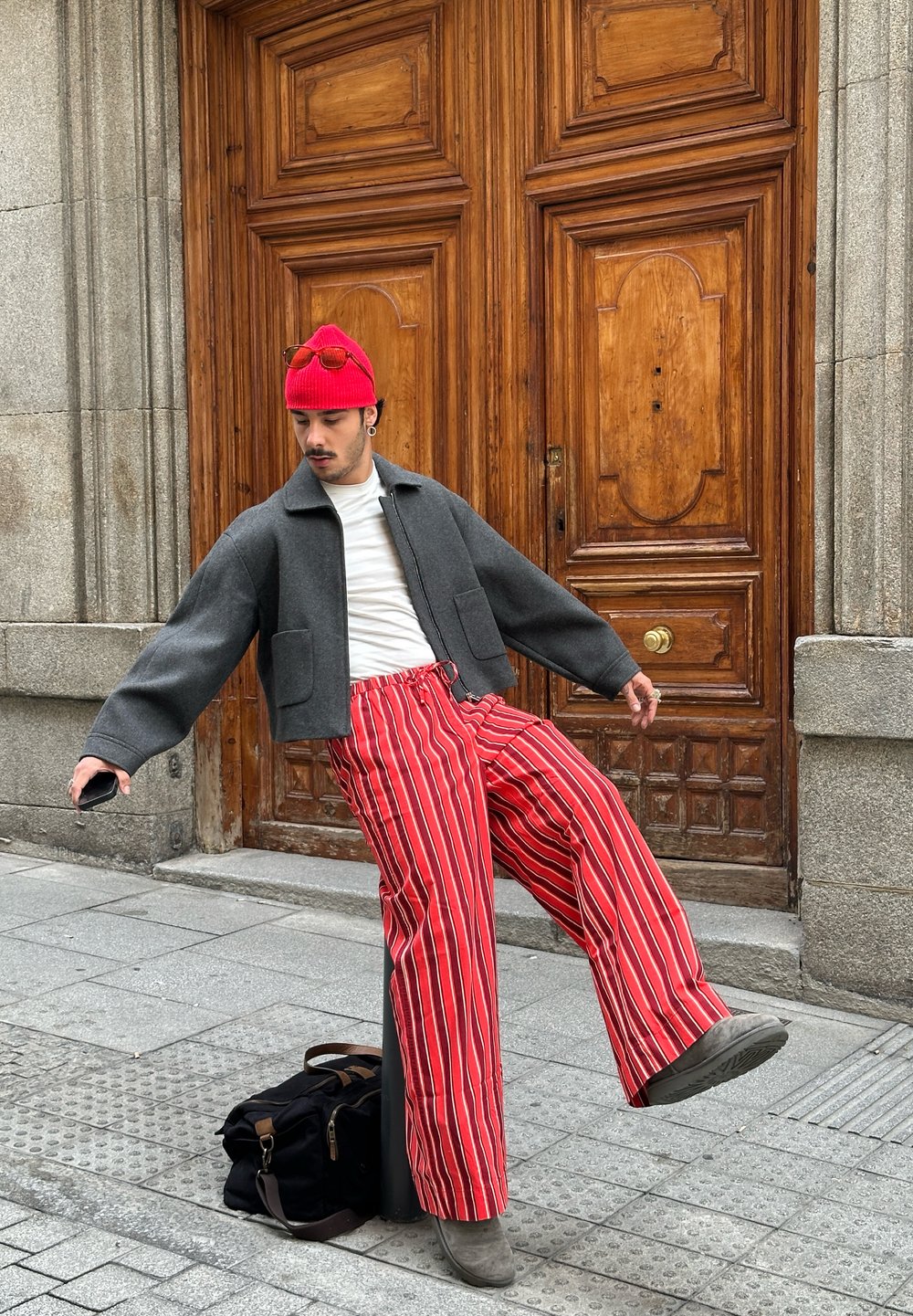 Γκρίζα κοντή ζακέτα, λευκό πουκάμισο, κόκκινα και άσπρα ριγέ παντελόνια με φαρδιά πόδια, γκρίζα υπόδηση, κόκκινο πλεκτό σκουφάκι, γυαλιά ηλίου, αστικό σκηνικό.