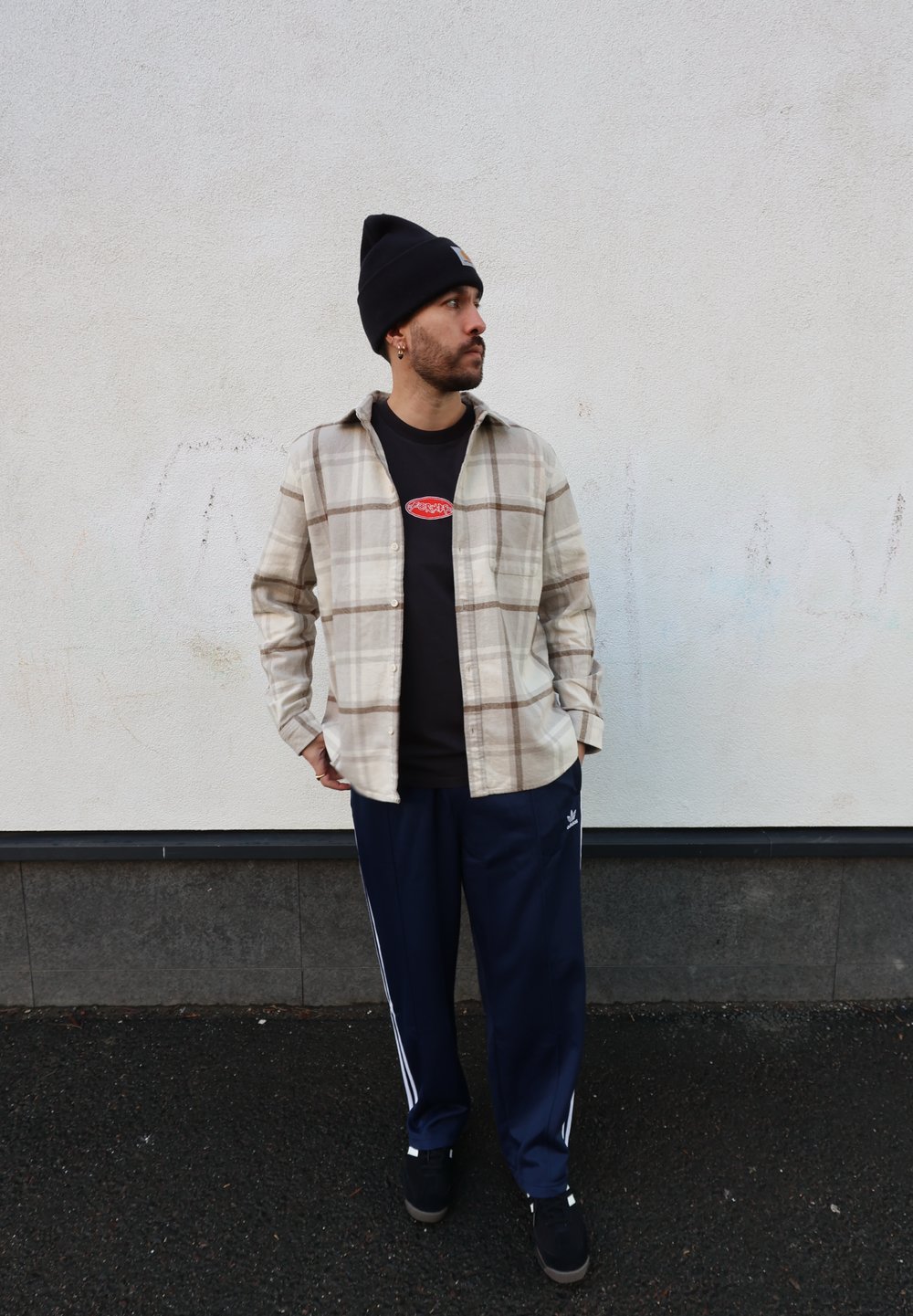 Chemise en flanelle à carreaux beige et marron, t-shirt noir avec logo, pantalon de survêtement bleu marine à rayures blanches, baskets noires et bonnet noir.