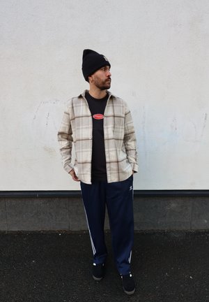 Camisa de franela a cuadros en beige y marrón, camiseta negra con logo, pantalones deportivos azul marino con rayas blancas, zapatillas negras y gorro negro.