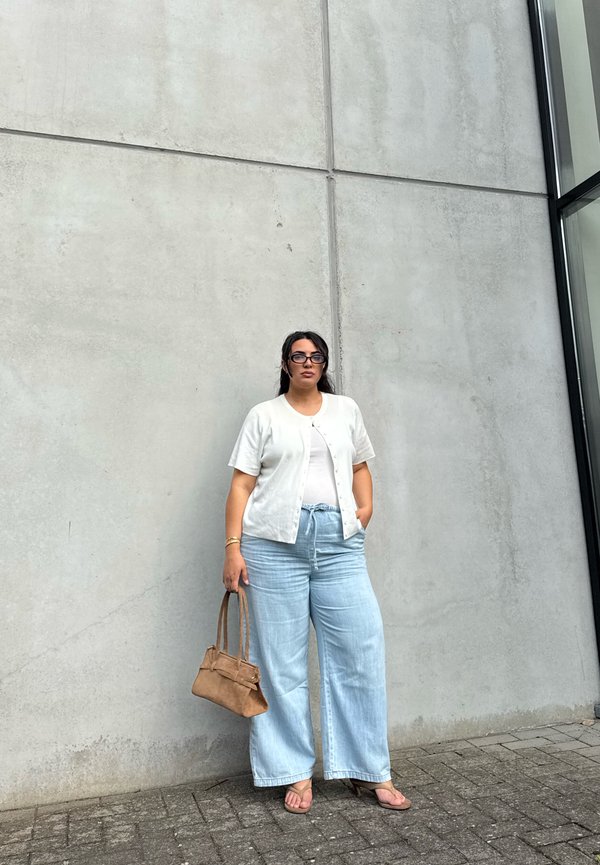 Mujer con cabello oscuro y gafas, de pie contra una pared de hormigón, lleva una blusa blanca, pantalones anchos azul claro y sostiene un bolso marrón.