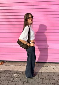 White cropped jumper, dark wide-leg jeans, and a brown shoulder bag. Standing against a pink striped wall, sunlight highlights textures.