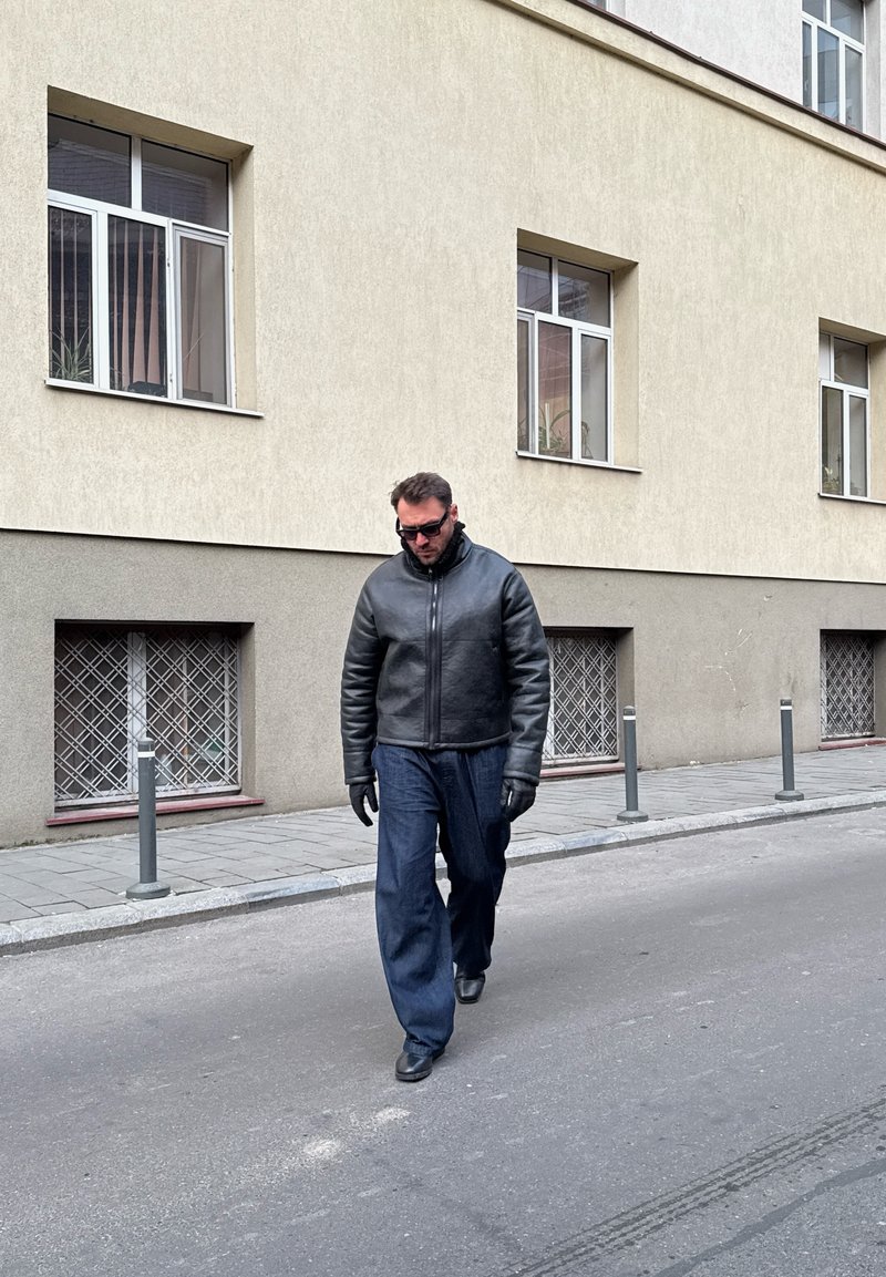 Mann, der dunkle Lederjacke, Handschuhe, Sonnenbrille und weite Jeans trägt, geht auf einer Straße neben einem beigen Gebäude mit vergitterten unteren Fenstern.