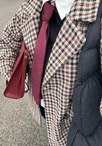 Brązowy i czarny płaszcz w pepitkę z dużymi klapami, bordo krawat oraz bordowa kopertówka, z warstwami czarnej bluzki i puchowej kamizelki.