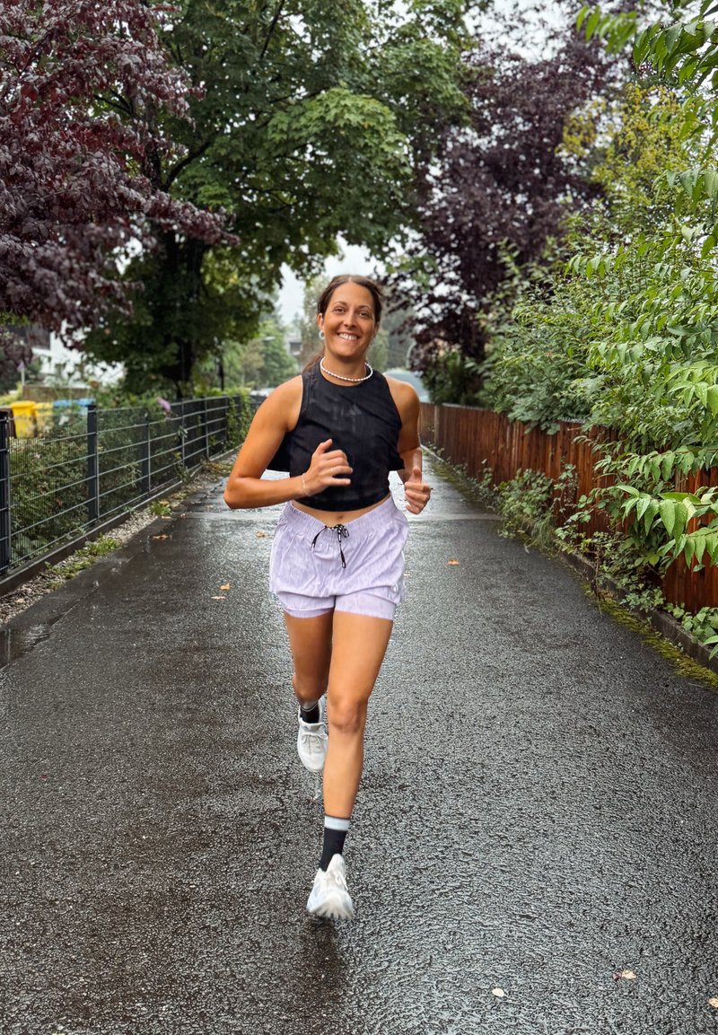 Donna che sorride mentre corre su un pavimento bagnato fiancheggiato da alberi e cespugli in una giornata nuvolosa.