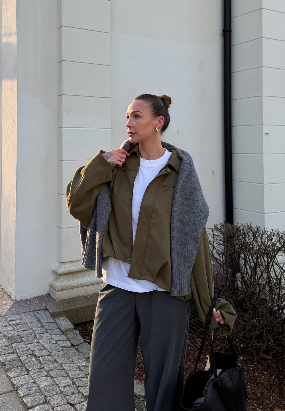 Chaqueta de ante verde oliva combinada con un suéter de punto gris, camiseta blanca debajo, pantalones anchos grises y un bolso negro. Materiales con textura presentes.