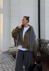 Chaqueta de ante verde oliva combinada con un suéter de punto gris, camiseta blanca debajo, pantalones anchos grises y un bolso negro. Materiales con textura presentes.