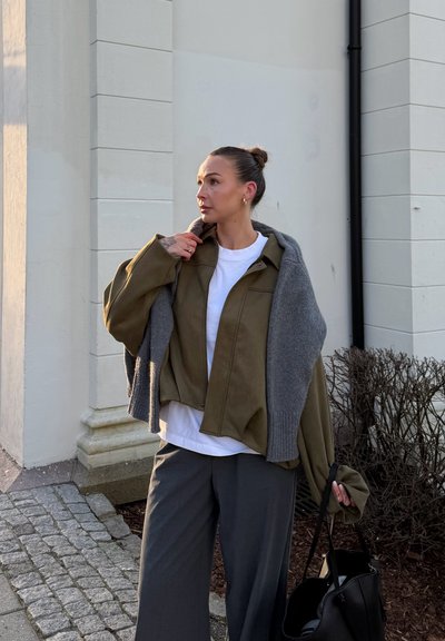 Chaqueta de ante verde oliva combinada con un suéter de punto gris, camiseta blanca debajo, pantalones anchos grises y un bolso negro. Materiales con textura presentes.