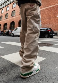 Personne portant un pantalon cargo beige et des baskets blanc-vert, debout sur un passage pour piétons dans une rue de la ville, avec des voitures garées et des bâtiments en brique à proximité.